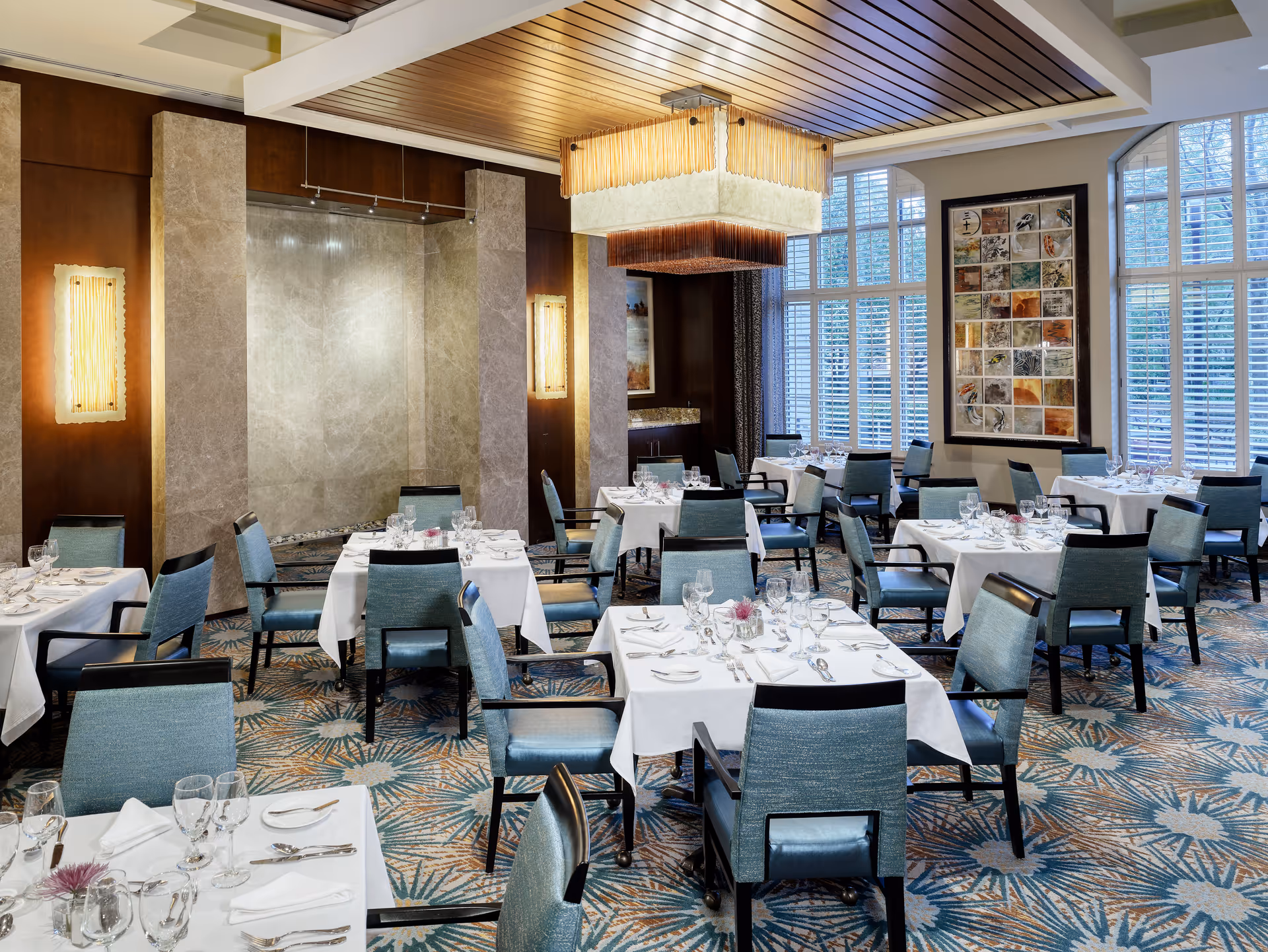 A well-lit dining room with multiple tables covered in white tablecloths, set with glassware, plates, and silverware. The room features blue upholstered chairs, large windows with white shutters, decorative wall lights, a modern chandelier, and a patterned carpet with blue and beige tones.