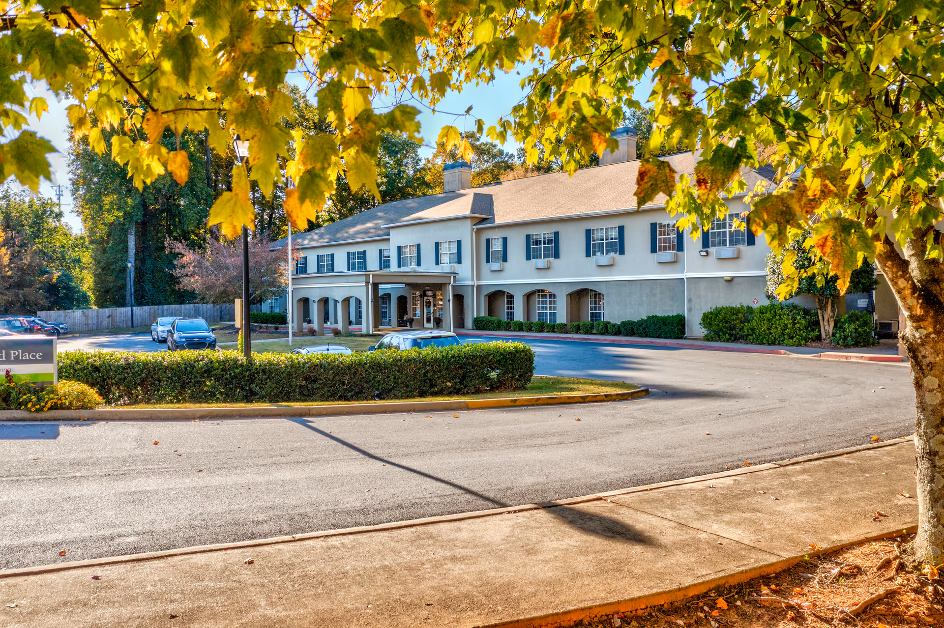 Exterior view of Greenwood Place Assisted Living and Memory Care building with a driveway and parking area in front. The building is two stories with multiple windows and a covered entrance. Trees with yellow and green leaves frame the image, indicating a fall season.