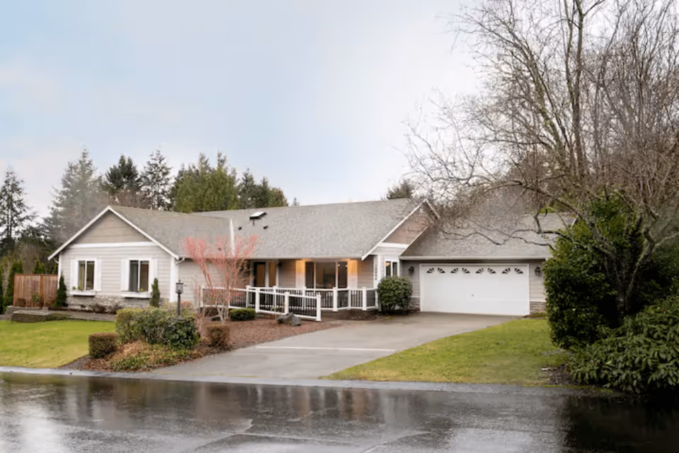 Single-story ranch-style house with an attached garage, front porch ramp, driveway and landscaped lawn on a rainy day.