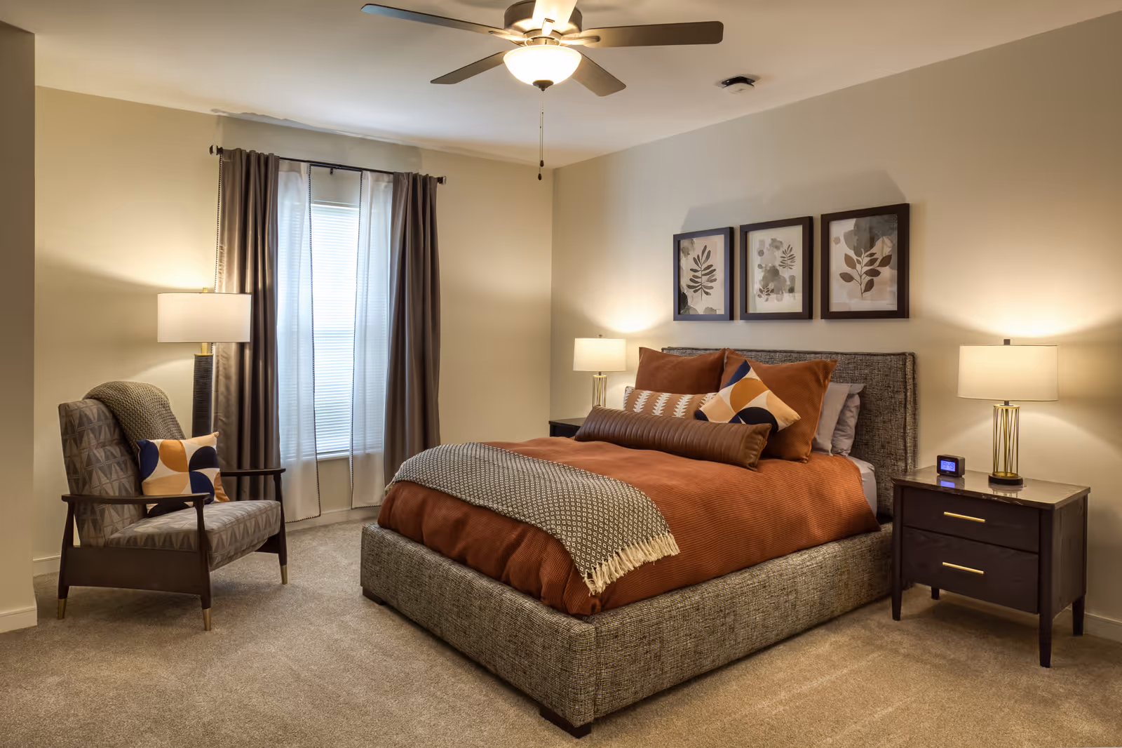 A cozy furnished bedroom with a large upholstered bed in rust bedding, two nightstands with lamps, an armchair, framed botanical prints, and a ceiling fan.