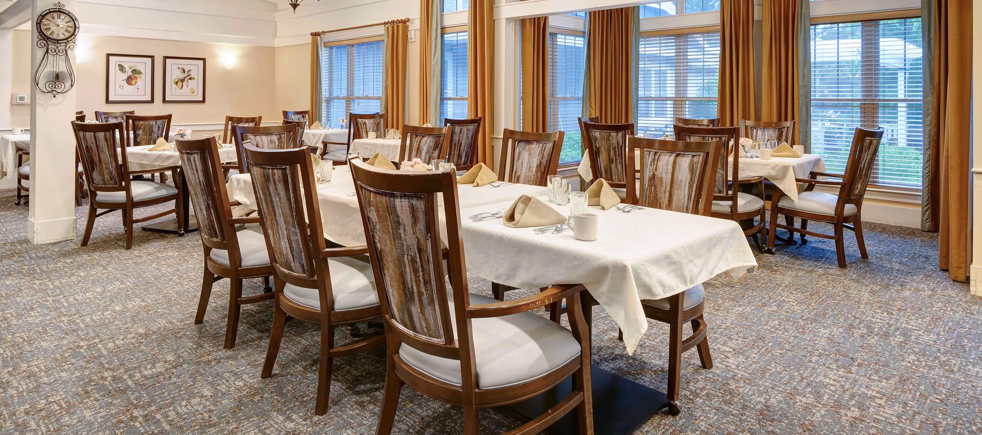A dining room with several wooden tables and chairs arranged neatly. The tables are covered with white tablecloths and set with beige napkins, glassware, and cups. Large windows with yellow curtains allow natural light to fill the room. The walls are decorated with framed botanical prints and a decorative wall clock.