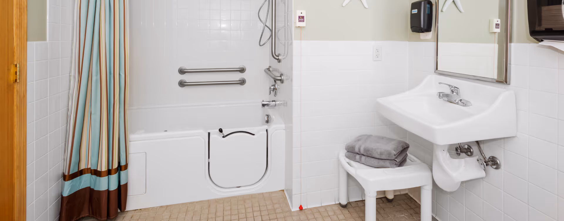 A bathroom with a walk-in bathtub featuring safety grab bars and a shower curtain. There is a white sink with a mirror above it, a soap dispenser mounted on the wall, and a small white stool with folded towels on it. The walls are tiled halfway up and the floor has small square tiles.