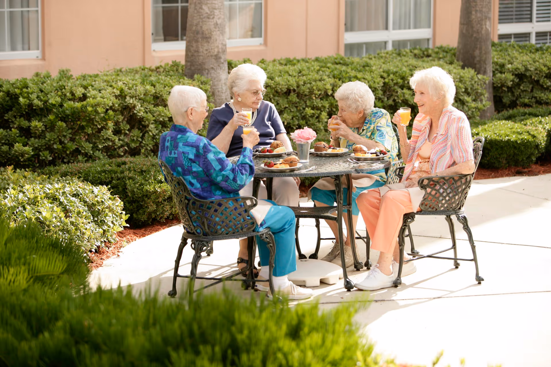 Four elderly women sitting around a metal outdoor table enjoying drinks and snacks in a garden area with green bushes and a building in the background.