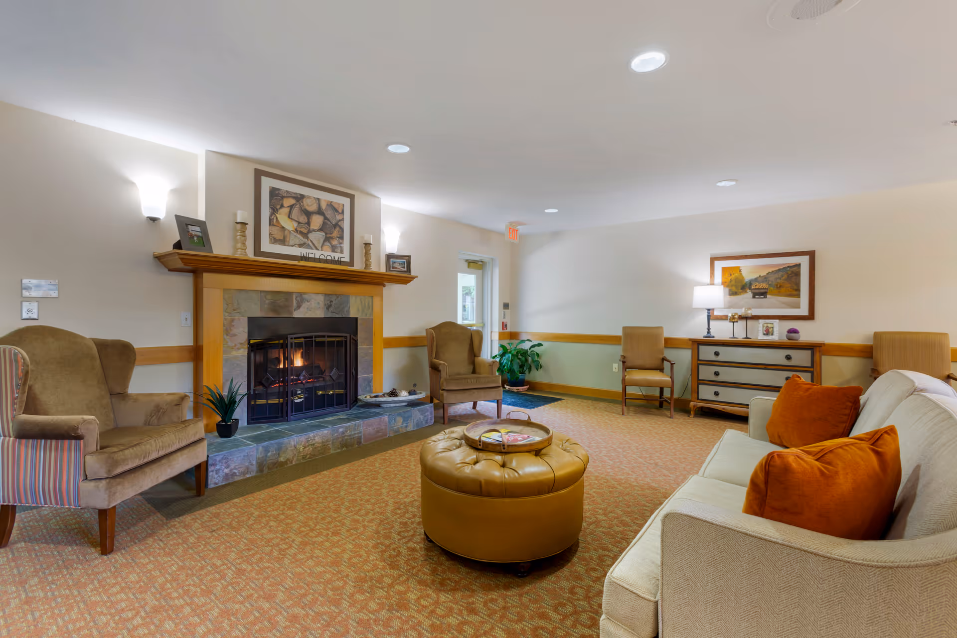 A cozy living room area with a lit fireplace surrounded by stone tiles and a wooden mantle. There are two armchairs on either side of the fireplace, a beige sofa with orange cushions, a round tan ottoman with a tray on top, a wooden dresser with a lamp and framed picture above it, and two additional chairs against the wall. The room has soft lighting and a carpeted floor with a patterned design.