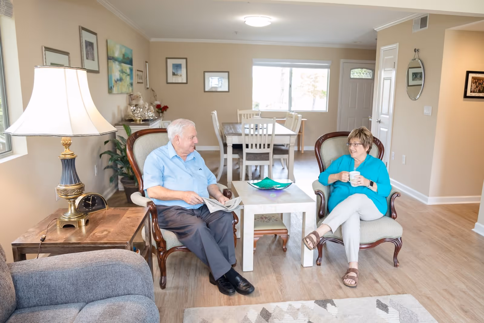 An elderly man and woman sitting in armchairs in a well-lit living room. The man is holding a newspaper and the woman is holding a mug. The room features a wooden side table with a lamp, a coffee table, a dining table with chairs in the background, and framed artwork on the walls.