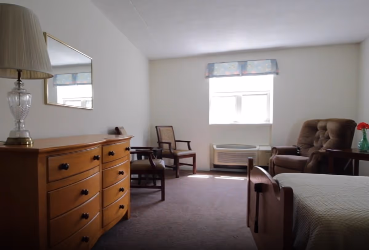 A simple, well-lit bedroom with a wooden dresser topped with a lamp and a mirror above it, two wooden chairs near a window with a floral valance, a brown upholstered armchair next to a small side table with a vase of red flowers, and a bed with a light-colored quilt.
