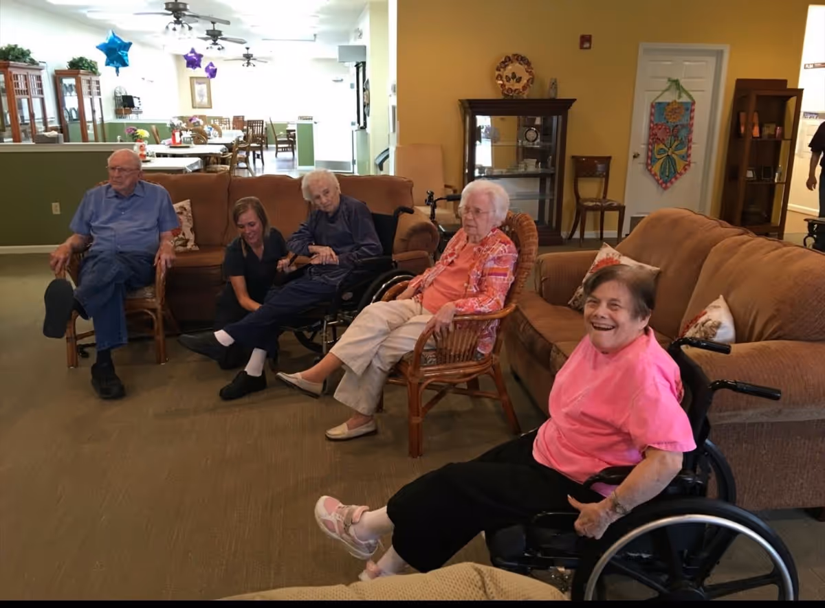 A group of elderly people sitting and relaxing in a cozy living room area of an assisted living facility. One woman in a pink shirt is smiling in a wheelchair, while others are seated on couches and chairs. A caregiver is kneeling beside one elderly woman in a wheelchair. The background shows a dining area with tables and chairs, decorated with star-shaped balloons.