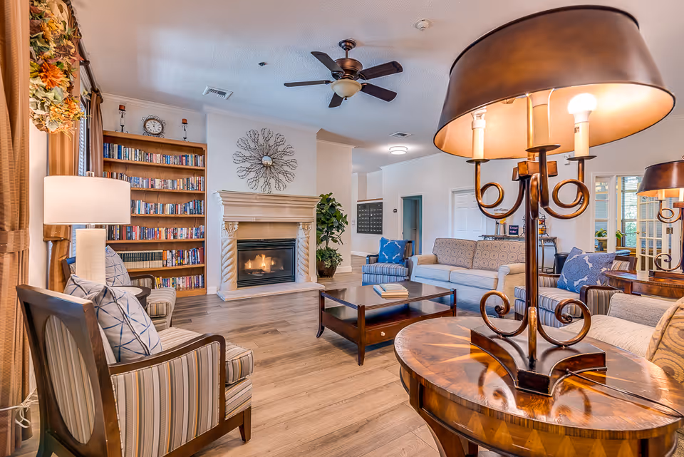 A cozy senior living facility common area with a lit fireplace, wooden bookshelves filled with books, comfortable armchairs and sofas with cushions, a wooden coffee table, and warm lighting from table lamps. The room has wooden flooring and a ceiling fan, creating a welcoming and relaxing atmosphere.