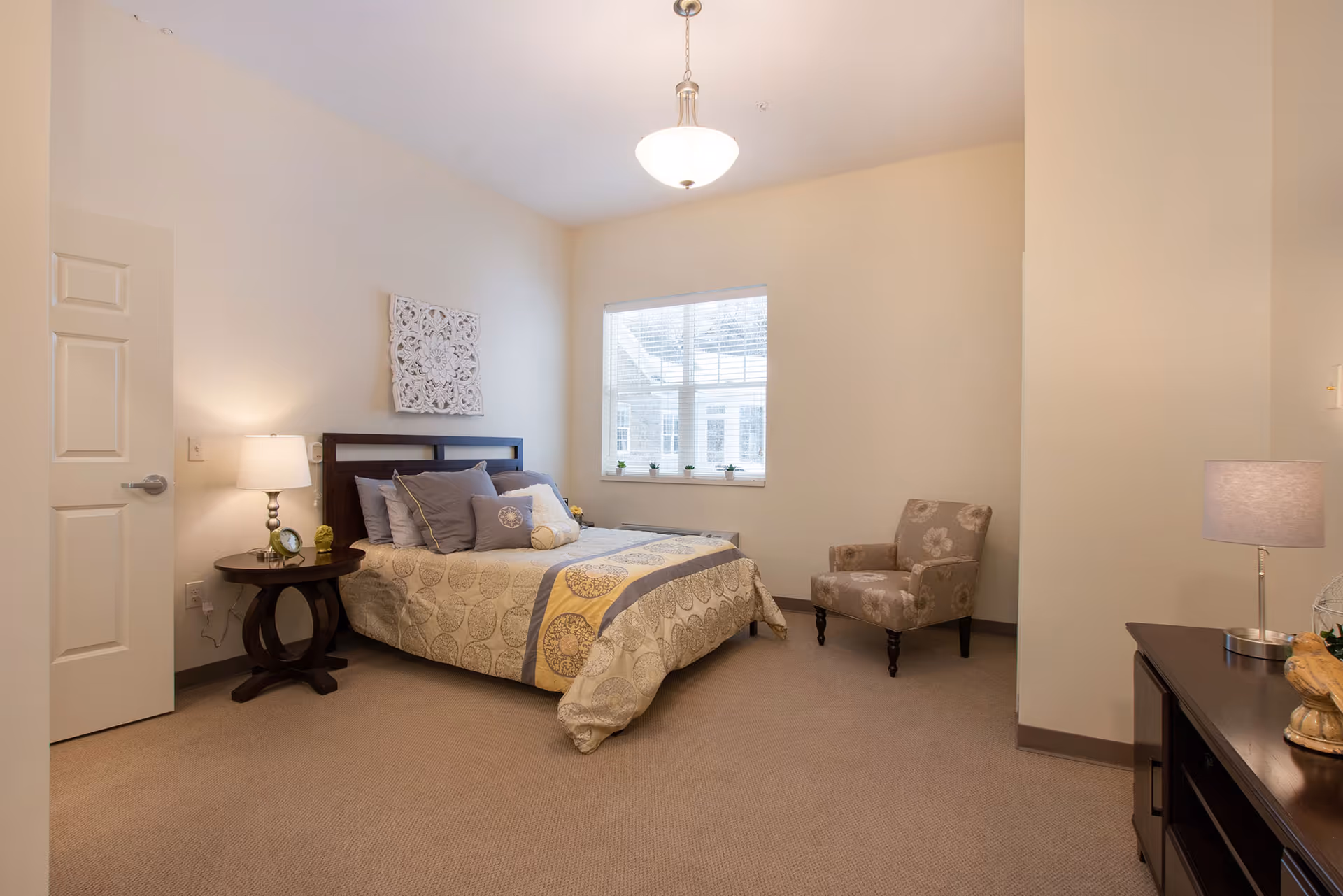 A cozy bedroom with a bed featuring a patterned bedspread and multiple pillows, a round wooden side table with a lamp and decorative items, a floral upholstered armchair, a window with blinds, and a dark wooden dresser with a lamp and decorative pieces.