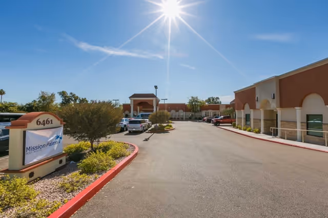 Front exterior of Mission Palms Post Acute with driveway, parked cars, landscaped median, and a bright sun overhead.