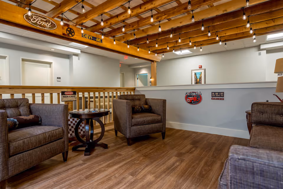 Cozy communal seating area with upholstered armchairs, a small round table, exposed wooden beams strung with lights, and vintage automotive signs on the wall.