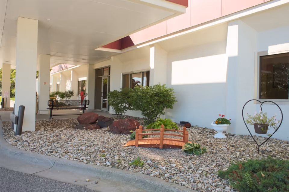Outdoor entrance area of a senior living facility with a covered walkway, a bench, small bushes, decorative rocks, a small wooden bridge, and a heart-shaped metal plant holder with a hanging flower pot.