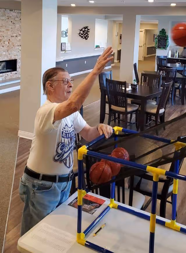 An elderly man wearing glasses and a white t-shirt is playing a tabletop basketball game inside a senior living facility. The background shows a dining area with tables and chairs, and a stone fireplace on the left side.