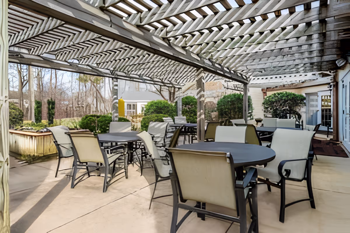 A shaded outdoor patio with round tables and multiple chairs under a wooden pergola beside landscaped shrubs.