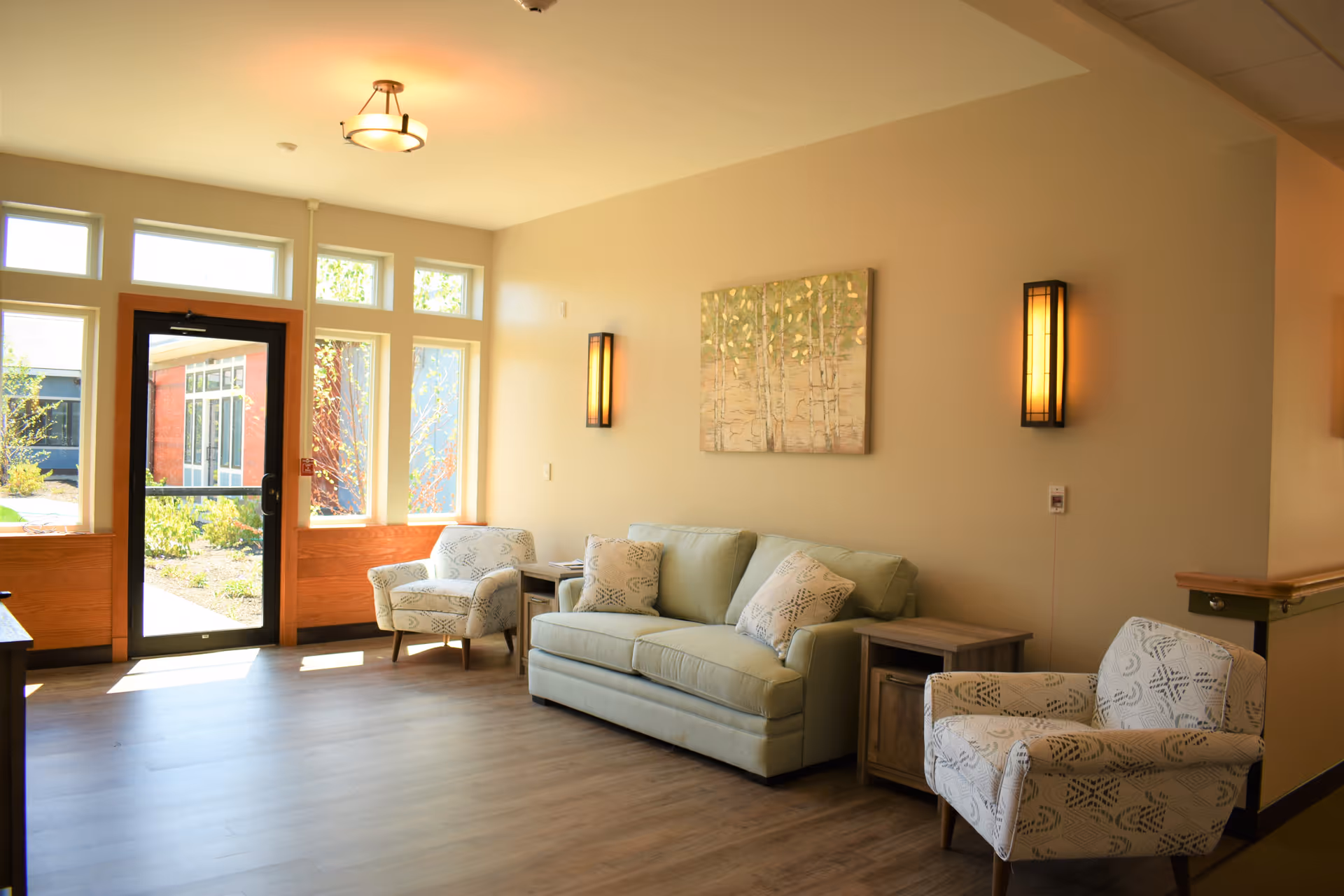 A bright and cozy living room area with a light green sofa, two patterned armchairs, wooden side tables, and a painting of trees on the wall. The room has large windows and a glass door letting in natural light, with warm wall sconces and a ceiling light fixture.