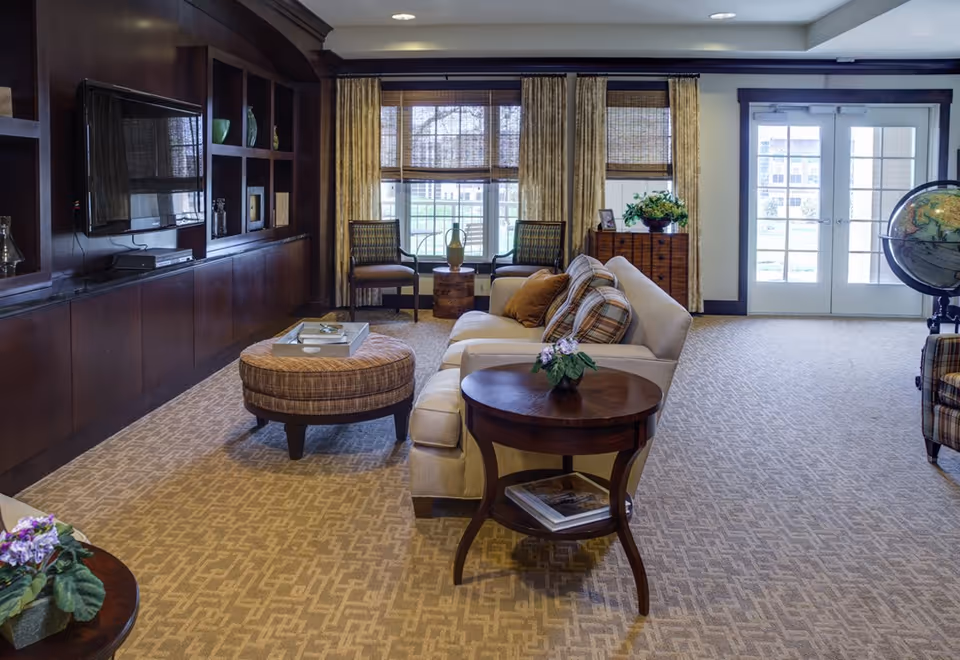 Bright, well-appointed common living room with sofas, an ottoman and side tables facing built-in shelving and a TV, with large windows and glass doors to the outside.