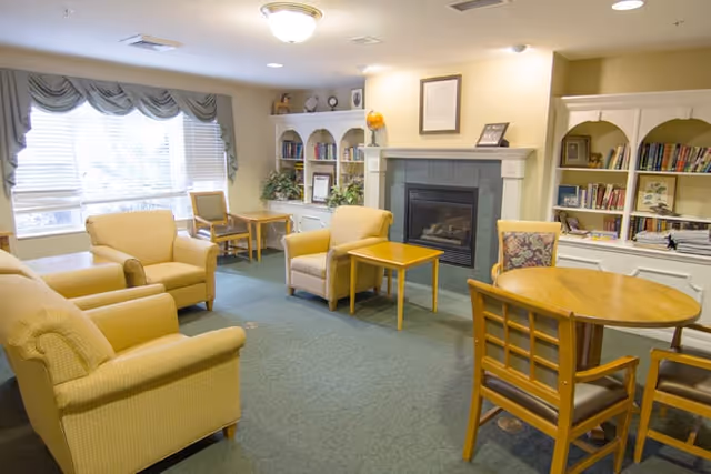 A cozy living room area with beige armchairs and wooden tables arranged around a fireplace. There are built-in white bookshelves filled with books and decorative items along the walls. A large window with draped curtains allows natural light to brighten the room.
