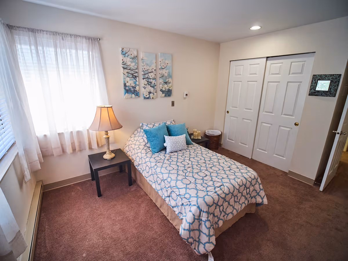 A cozy bedroom with a single bed covered in a white and blue patterned bedspread and several blue and white pillows. There is a black nightstand with a lamp beside the bed. The room has beige walls, a window with sheer white curtains, a brown carpet, and a closet with white sliding doors. Three decorative panels with a floral design hang above the bed.