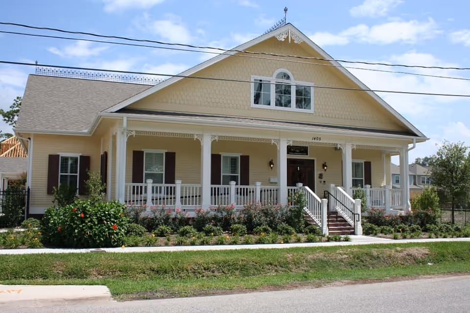 Front exterior view of a yellow single-story building with white trim and a covered porch with white railings. There are steps leading up to the entrance door, surrounded by bushes and flowers, under a partly cloudy blue sky.