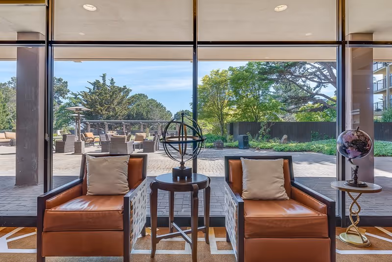 Two brown leather armchairs with beige cushions are positioned on either side of a small round wooden table with a decorative armillary sphere on it. To the right, there is a small round side table with a globe on top. Large floor-to-ceiling windows behind the chairs provide a clear view of an outdoor patio area with additional seating and greenery, including trees and shrubs under a blue sky.