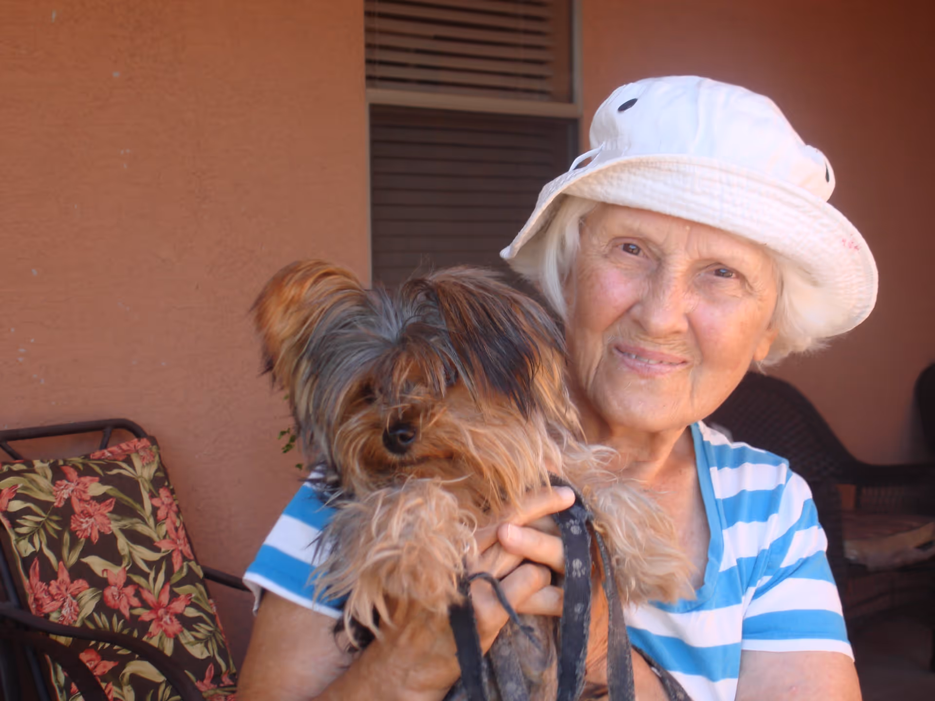 An elderly woman wearing a white hat and a blue and white striped shirt is holding a small fluffy dog. They are sitting outside on a patio with a brown wall and a chair with a floral cushion in the background.