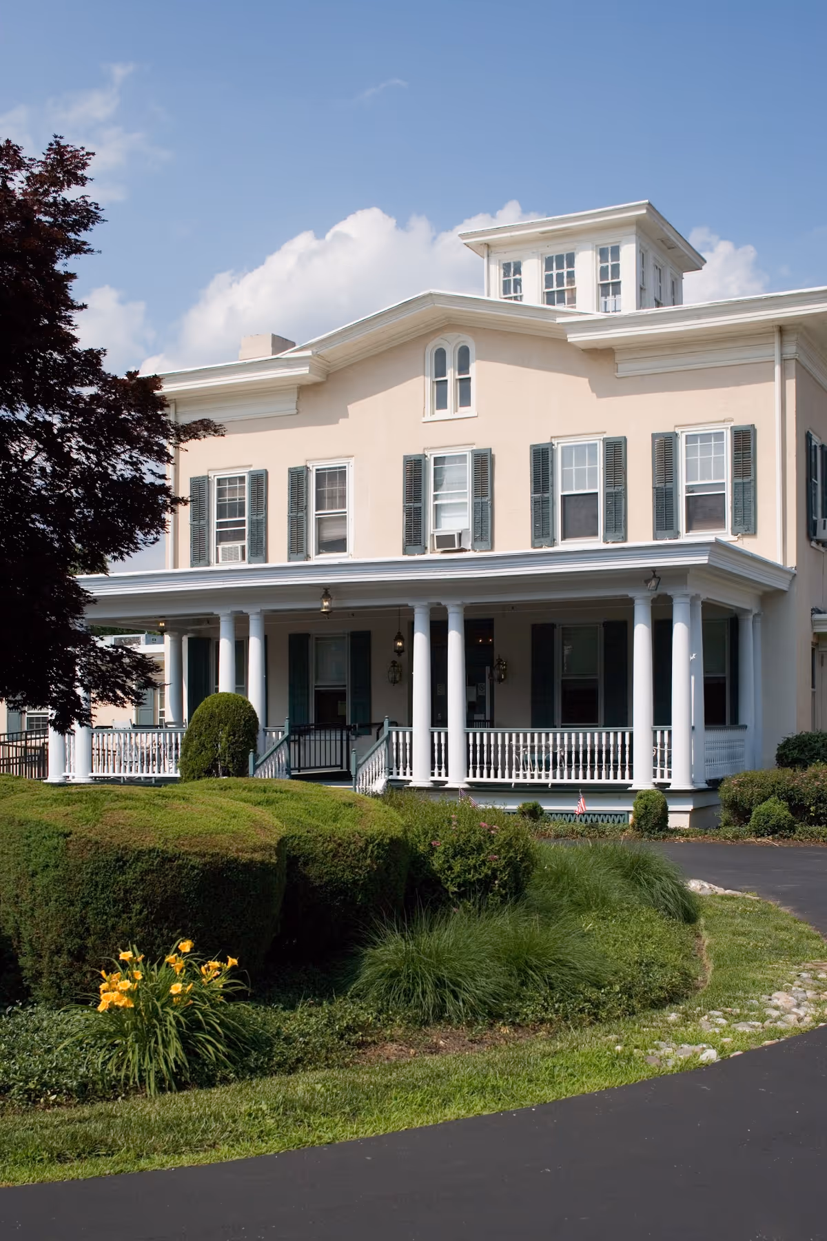 A large, light-colored two-story building with green shutters and a wraparound porch supported by white columns. The building is surrounded by neatly trimmed bushes, green grass, and a paved driveway. The sky is clear with a few clouds.