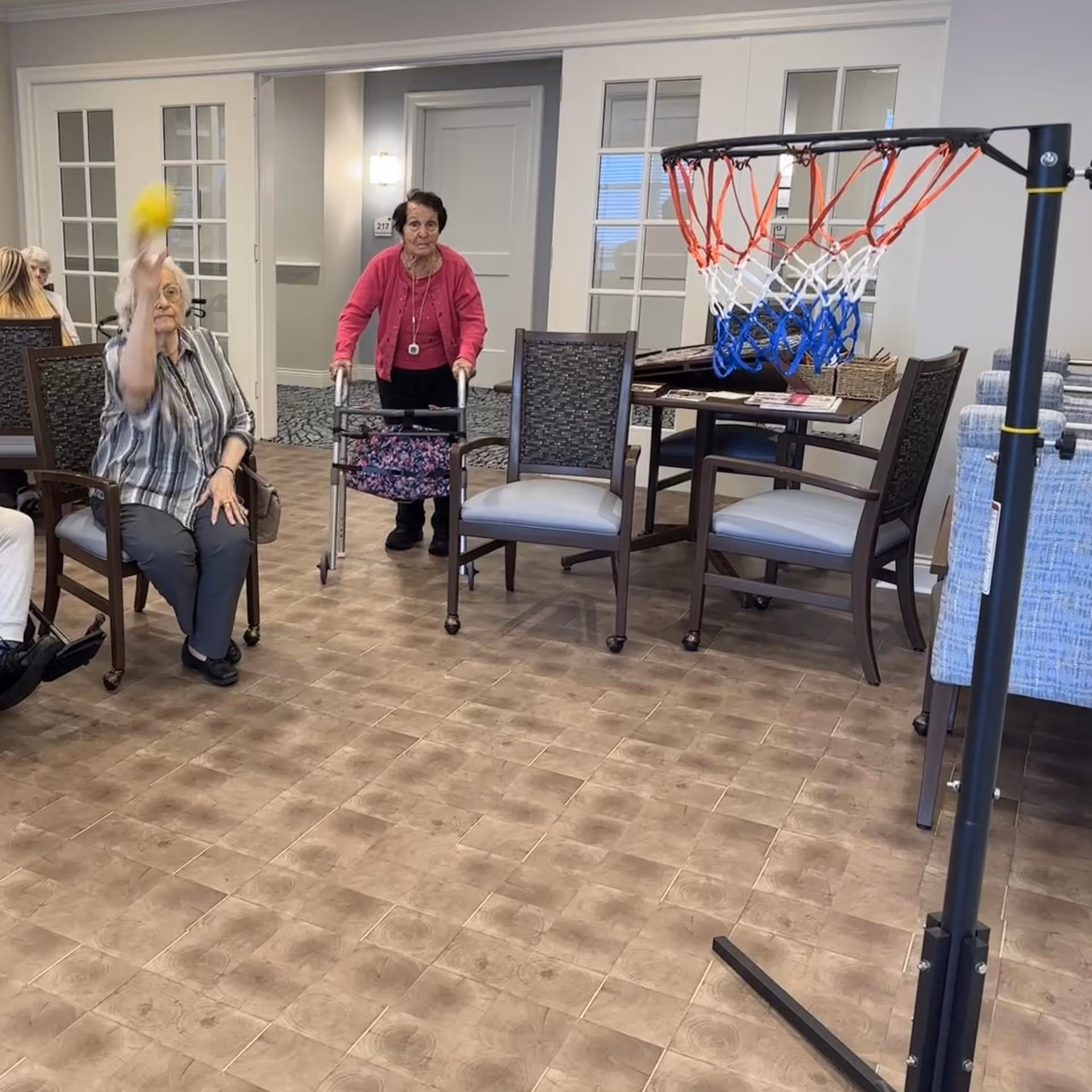 Two elderly women in a common area of a senior living facility. One woman is seated and throwing a yellow ball towards a small basketball hoop, while the other woman stands behind her using a walker. The room has chairs, a table with magazines, and a door labeled 217 in the background.
