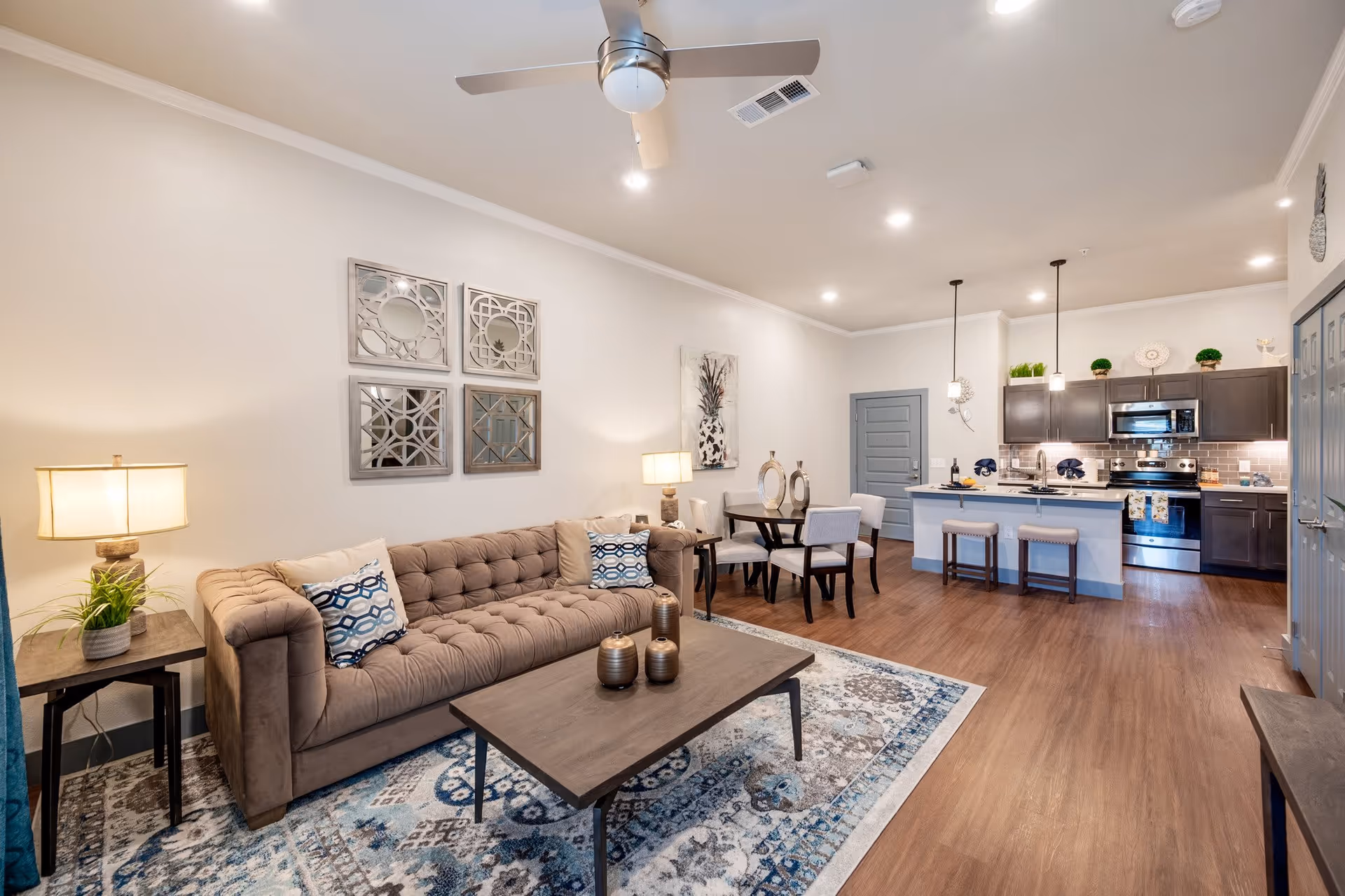 A modern and cozy living room and kitchen area in a senior living facility. The living room features a beige tufted sofa with patterned cushions, a wooden coffee table with decorative vases, a side table with a lamp and plant, and a patterned area rug. The kitchen has dark cabinets, stainless steel appliances, a breakfast bar with two stools, and pendant lights hanging above. A small dining table with four chairs is situated between the living room and kitchen. The walls are light-colored with decorative mirrors and artwork, and the floor is wood.