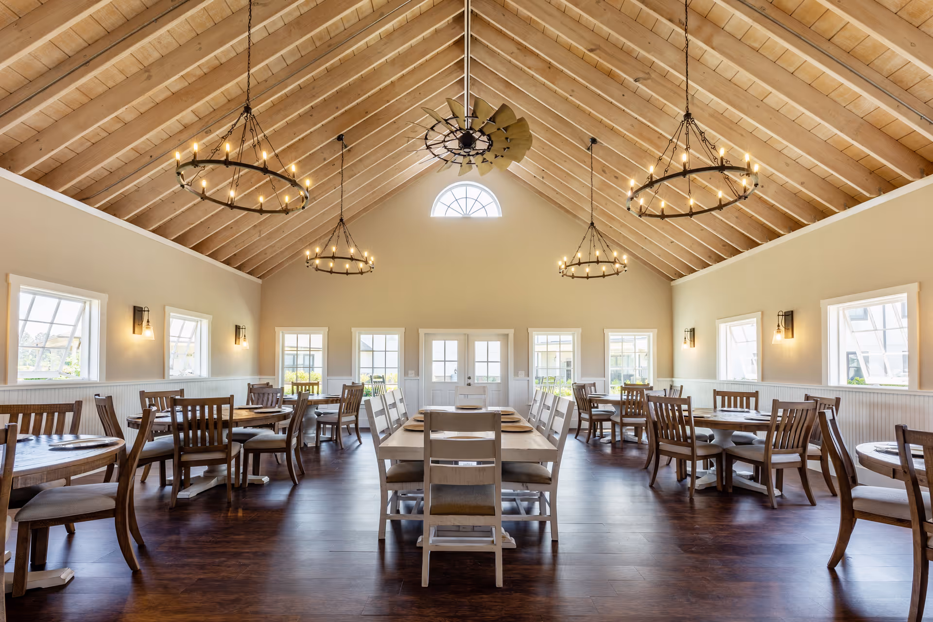 A spacious dining room with high vaulted wooden ceilings and large windows letting in natural light. The room features multiple wooden dining tables and chairs arranged neatly on a dark wood floor. Three large circular chandeliers hang from the ceiling, and a decorative ceiling fan is mounted in the center. The walls are painted a soft beige with white wainscoting and wall sconces providing additional lighting.