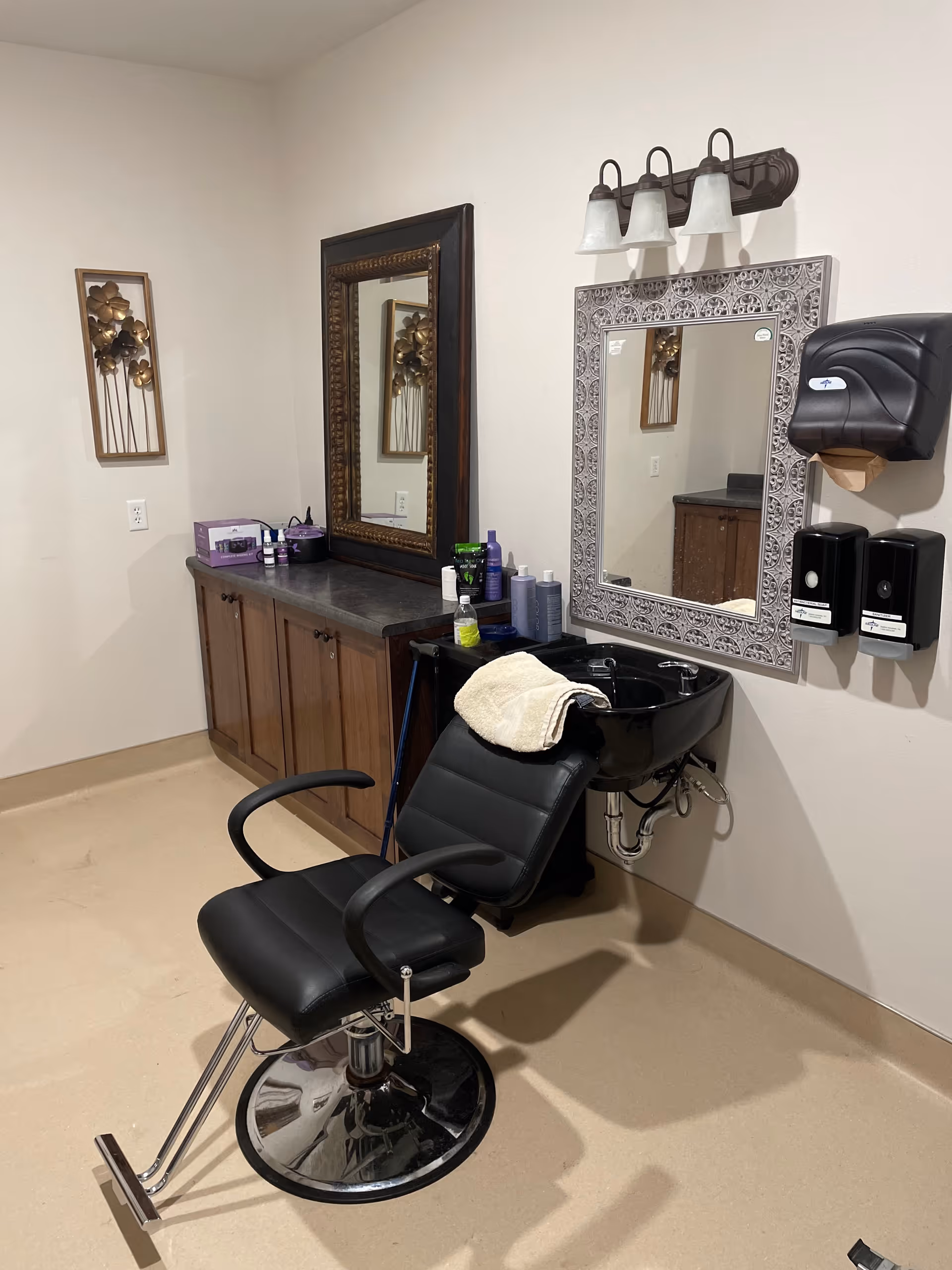 A salon area with a black reclining chair in front of a black wash basin. There are two mirrors on the wall, one with a decorative frame and the other with a dark wooden frame. Below the mirrors is a countertop with wooden cabinets and various hair care products. On the wall next to the wash basin are mounted dispensers and a paper towel holder. The room has beige flooring and light-colored walls.