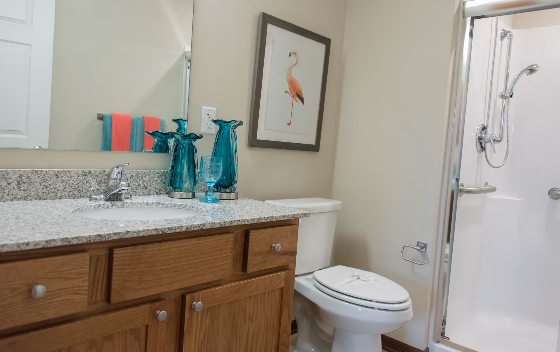A bathroom with a wooden vanity topped with a granite countertop, a sink with a chrome faucet, and decorative blue glass vases and a glass. A toilet is next to the vanity, and a framed picture of a flamingo hangs on the wall above it. A shower with a glass door and chrome fixtures is visible on the right side. Two colorful towels hang on a rack reflected in the mirror.