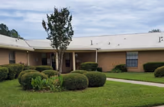 Single-story brick building with a manicured lawn, round shrubs, a sidewalk, and a cloudy sky.