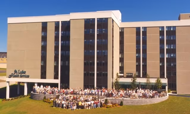 Large group of people gathered outside in front of a multi-story building with the sign 'St. Luke Health Services' on the left side. The building has many windows and a clear blue sky above.