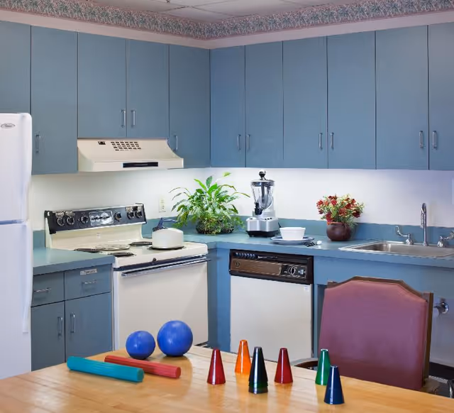 A kitchen area with blue cabinets, a white refrigerator, stove, and dishwasher. There is a blender, a potted plant, and a vase with flowers on the countertop. In the foreground, a wooden table holds two blue balls, two foam rollers, and several colorful cone-shaped objects. A maroon chair is positioned at the table.