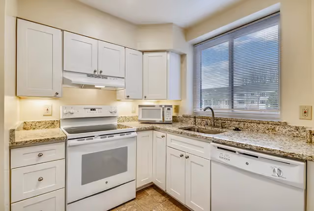 A clean and modern kitchen featuring white cabinets, a white electric stove with an overhead range hood, a microwave on the countertop, a stainless steel sink under a window with blinds, and a white dishwasher. The countertops are granite, and the floor has brown tiles.