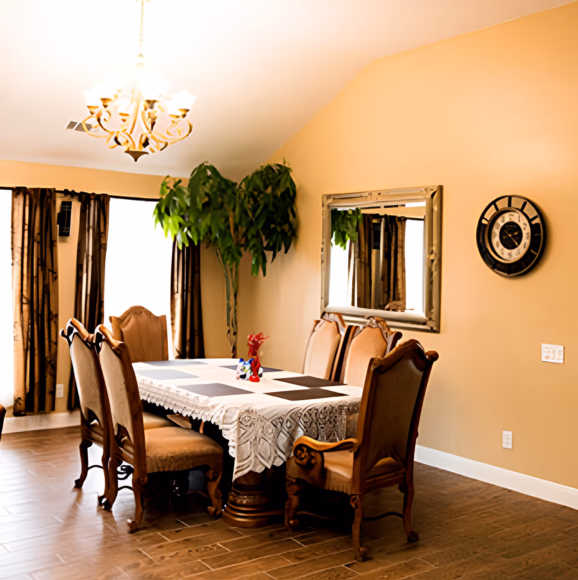 A dining room with a rectangular table covered by a white lace tablecloth and black placemats. Six upholstered wooden chairs surround the table. A large green potted plant is in the corner near windows with dark curtains. A decorative wall clock and a large mirror hang on the beige wall. A chandelier with multiple lights hangs from the ceiling.