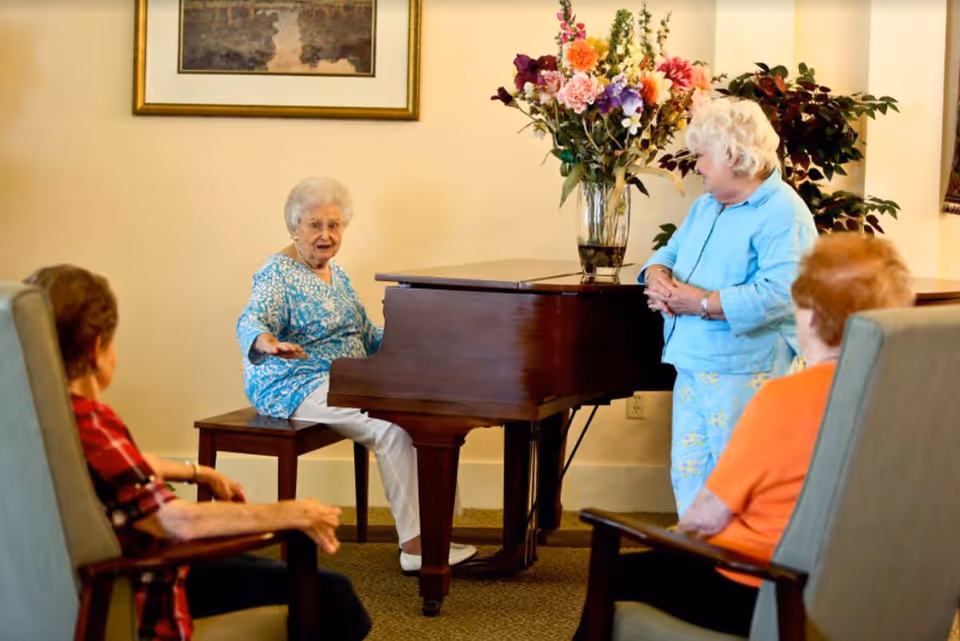 Four elderly women in a common room at Azalea Estates of Gonzales. One woman is seated at a grand piano, appearing to play or speak, while another woman stands nearby smiling. Two other women sit in armchairs facing the piano. A large vase of colorful flowers is on top of the piano, and a framed painting hangs on the wall behind them.