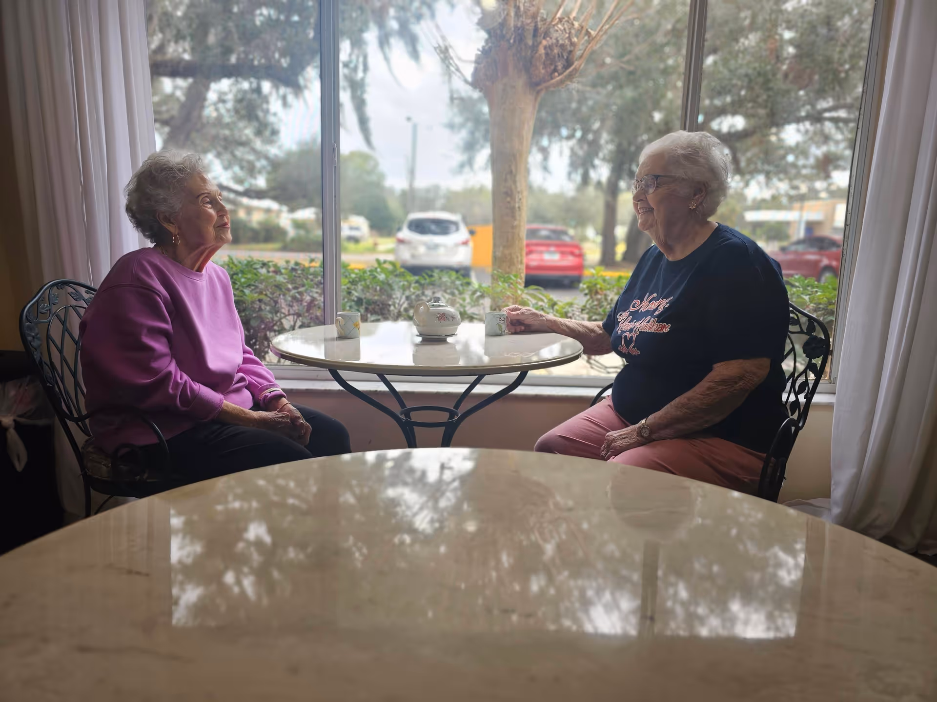 Two elderly women sitting at a round table near a large window with a view of trees and parked cars outside. They are engaged in conversation, each with a cup on the table. One woman is wearing a purple sweater and the other a dark shirt with pink pants.