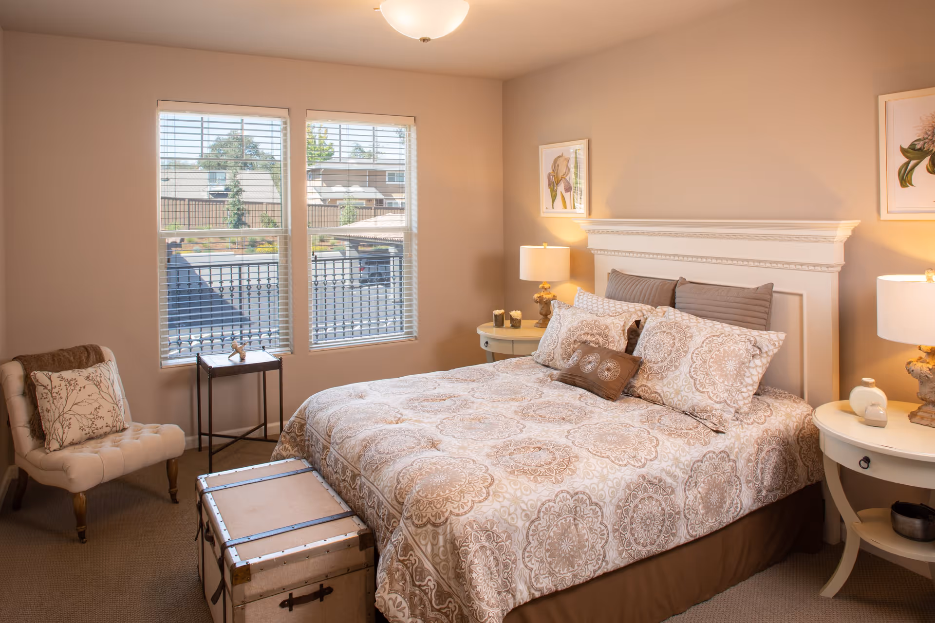 A cozy bedroom with a large bed featuring patterned bedding and multiple pillows. There are two round bedside tables with lamps on each side of the bed. A cushioned chair with a decorative pillow is placed near two windows with blinds, and a small side table is positioned between the chair and the windows. The walls are adorned with framed floral artwork, and a trunk is situated at the foot of the bed.