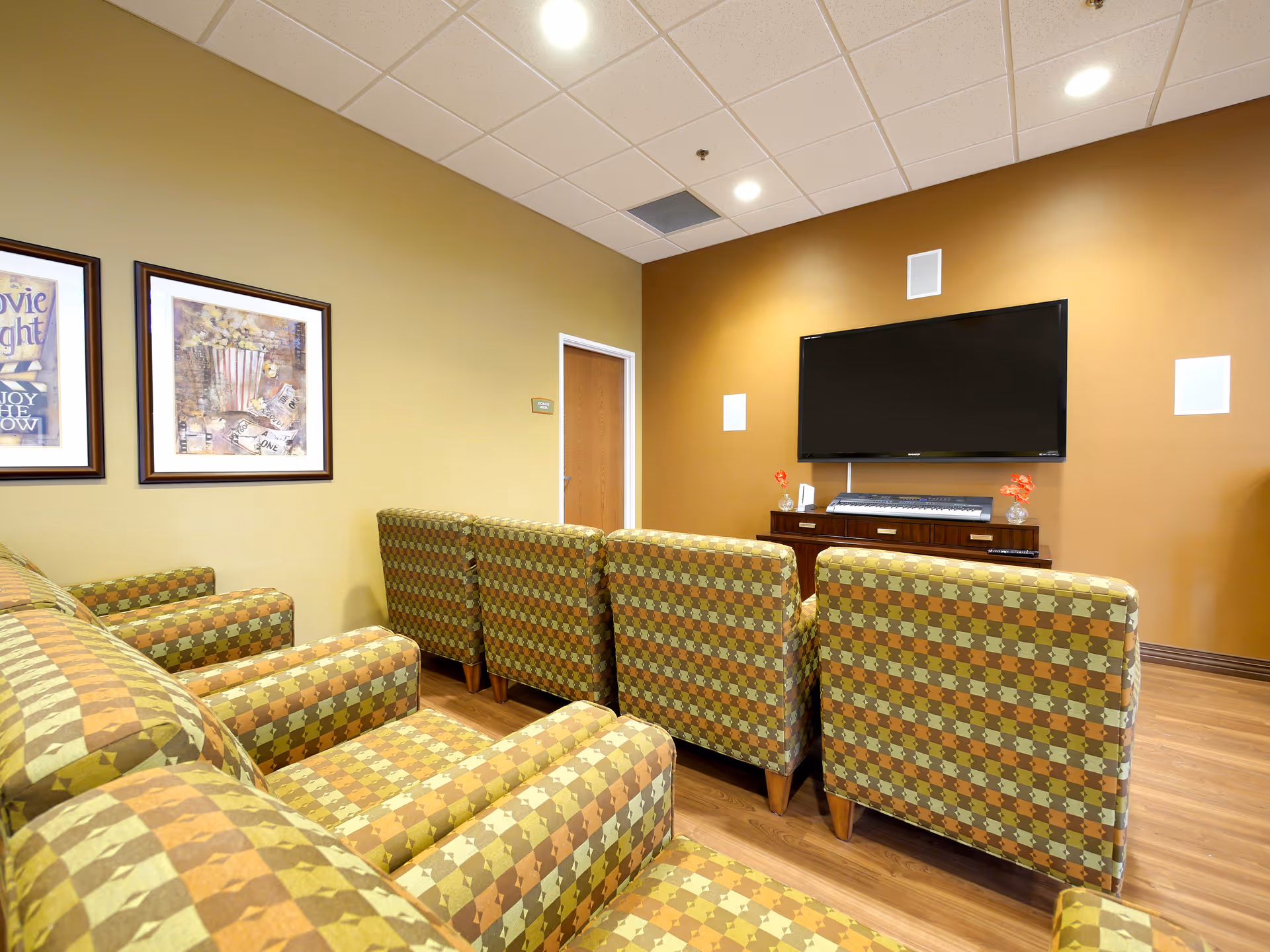 A cozy living room area with patterned armchairs and a sofa arranged facing a large flat-screen TV mounted on a brown accent wall. The room has wood flooring, recessed ceiling lights, and two framed pictures on a beige wall.