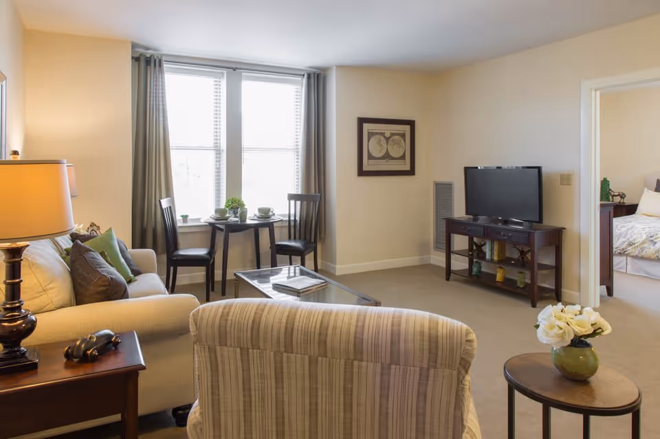 A cozy living room with a beige sofa, a striped armchair, a wooden side table with a lamp, and a small round table with two chairs near a window with curtains. A TV is placed on a wooden stand against the wall, and a doorway leads to a bedroom with a bed and nightstand visible. A small round table with a green vase and white flowers is in the foreground.