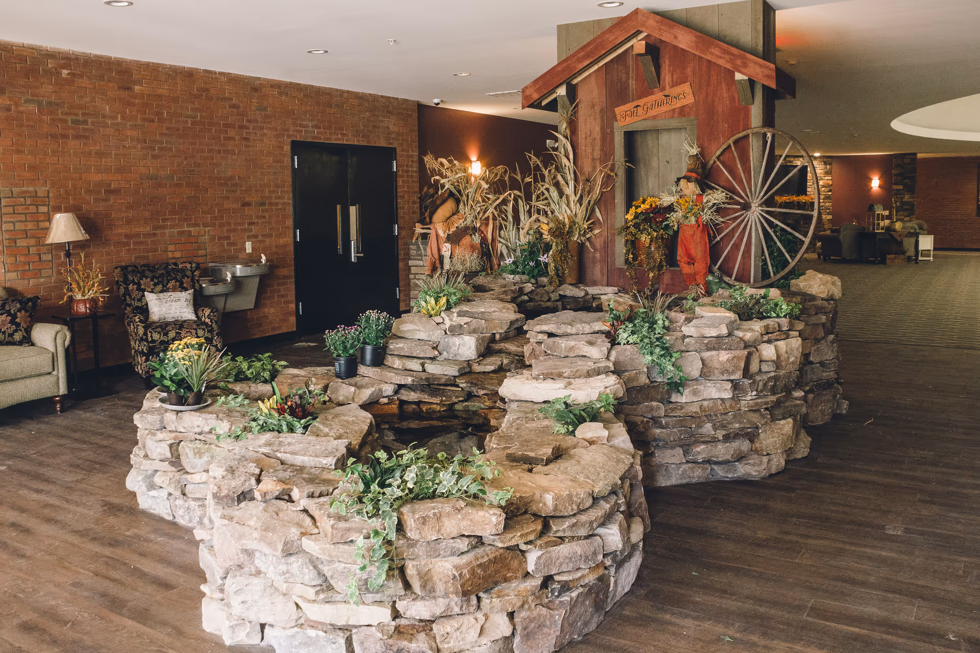Indoor lobby area with a tiered stone planter/fountain, seasonal decorations including a wagon wheel, and seating along a brick wall.
