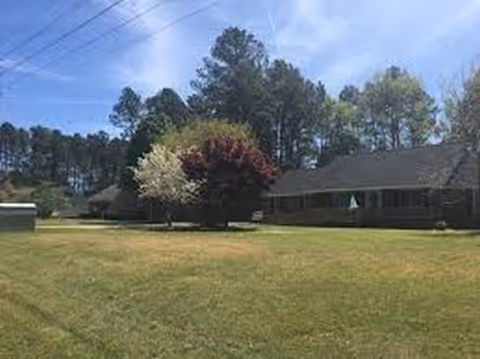 Single-story care facility with a grassy lawn, trees, and a front entrance under a blue sky.