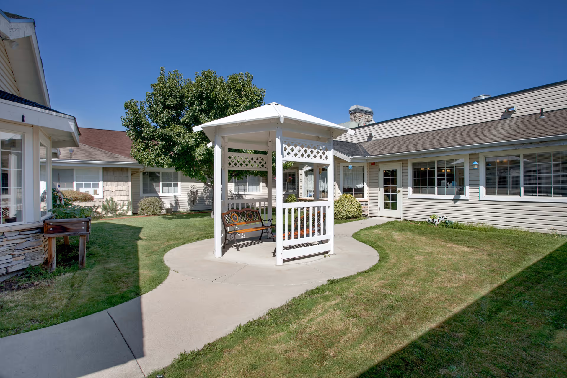 Outdoor courtyard area with a white gazebo featuring a bench swing, surrounded by a concrete pathway and green grass. The courtyard is enclosed by single-story buildings with beige siding and multiple windows under a clear blue sky.