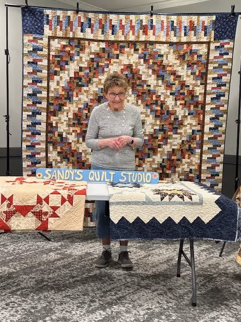 An elderly woman standing behind a table displaying two quilts in front of a large, colorful quilt hanging on the wall. A sign on the table reads 'SANDY'S QUILT STUDIO'. The setting appears to be an indoor room with a carpeted floor.