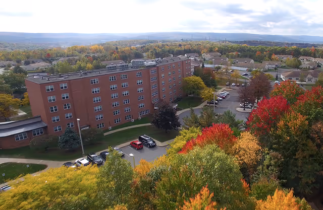 Aerial view of a multi-story brick building surrounded by colorful autumn trees with a parking lot and residential houses in the background under a cloudy sky.