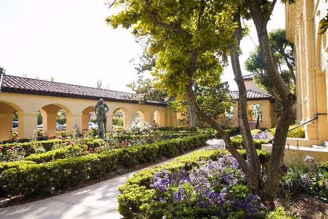 A sunny outdoor garden area at Hollenbeck Palms featuring a paved walkway, neatly trimmed hedges, blooming purple and pink flowers, several trees providing shade, and a statue of two figures near an arched colonnade structure with a tiled roof.
