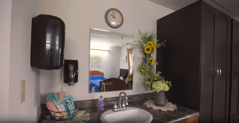 A bathroom sink area with a mirror above it reflecting a bedroom with a bed and a TV. The sink countertop has a soap dispenser, a basket with towels and toiletries, and a vase with sunflowers and other flowers. A wall-mounted black paper towel dispenser is on the left side of the mirror, and a clock is mounted above the mirror.