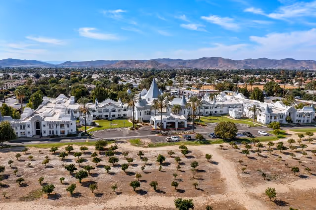 Aerial view of Camelot - A Provincial Senior Living Community, featuring a large white building with castle-like turrets and towers surrounded by palm trees and greenery, with a dry landscape and small trees in the foreground and mountains in the background.