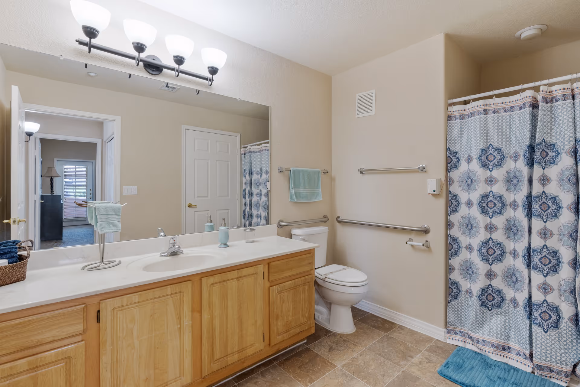 A clean and well-lit bathroom with a large mirror above a double sink vanity with wooden cabinets. There is a toilet with grab bars on the wall beside it, a shower with a blue and white patterned curtain, and a blue bath mat on the tiled floor. A towel rack with a green towel is mounted on the wall, and a door leads to another room visible through the mirror.